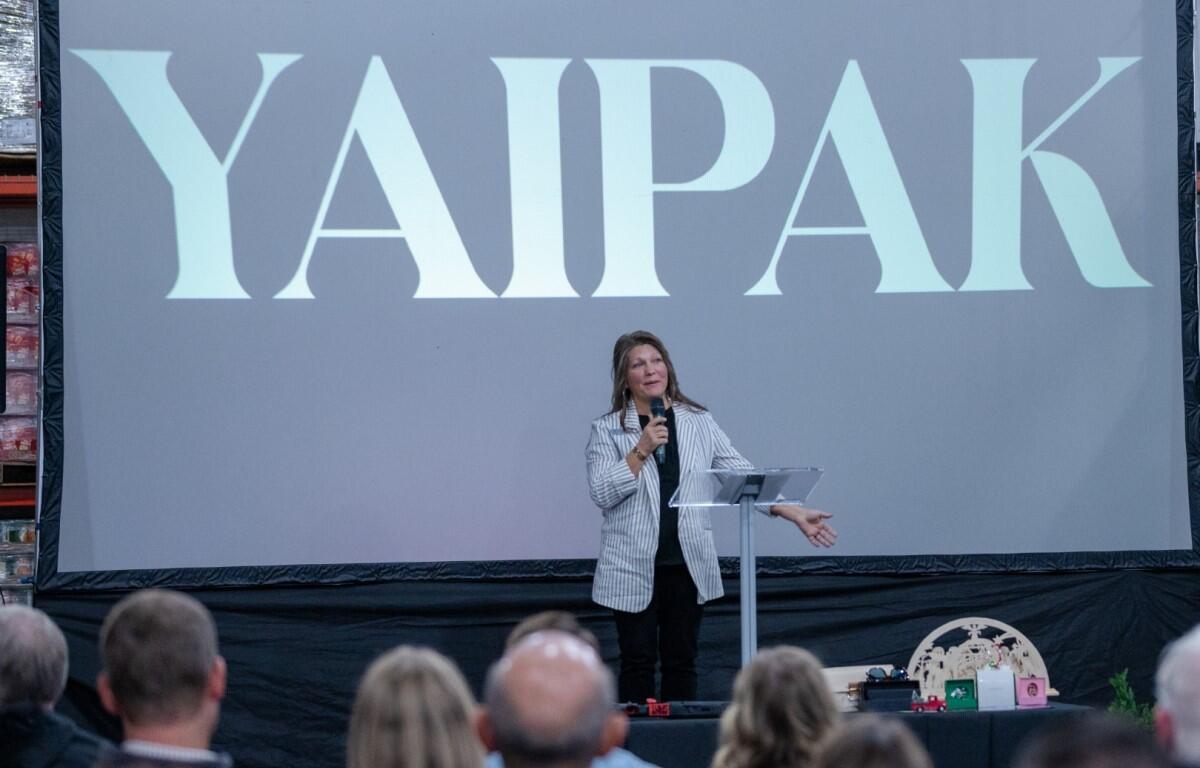 Woman in a striped blazer speaks at a podium on a stage, a large screen behind reads 'YAPAK', audience in the foreground.