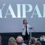Woman in a striped blazer speaks at a podium on a stage, a large screen behind reads 'YAPAK', audience in the foreground.