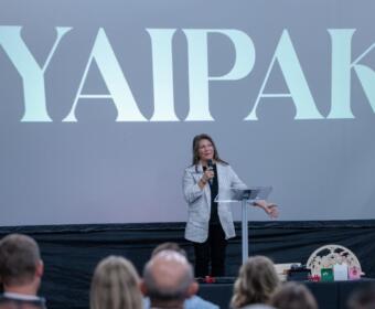Woman in a striped blazer speaks at a podium on a stage, a large screen behind reads 'YAPAK', audience in the foreground.