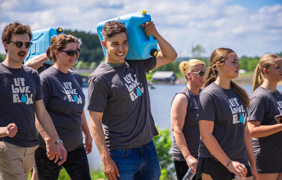 Group of volunteers in matching Let Love Flow shirts walking outdoors by a lake, carrying blue water containers.
