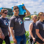 Group of volunteers in matching Let Love Flow shirts walking outdoors by a lake, carrying blue water containers.