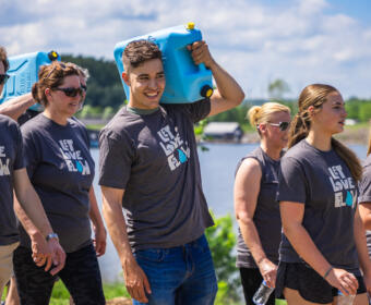 Group of volunteers in matching Let Love Flow shirts walking outdoors by a lake, carrying blue water containers.