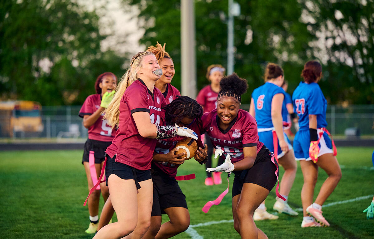 A group of girls in maroon jerseys tackle for the football during a flag football game on a green field, pink flag belts fluttering nearby.