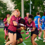 A group of girls in maroon jerseys tackle for the football during a flag football game on a green field, pink flag belts fluttering nearby.