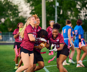 A group of girls in maroon jerseys tackle for the football during a flag football game on a green field, pink flag belts fluttering nearby.