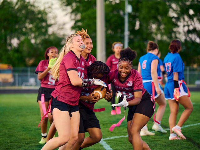 A group of girls in maroon jerseys tackle for the football during a flag football game on a green field, pink flag belts fluttering nearby.