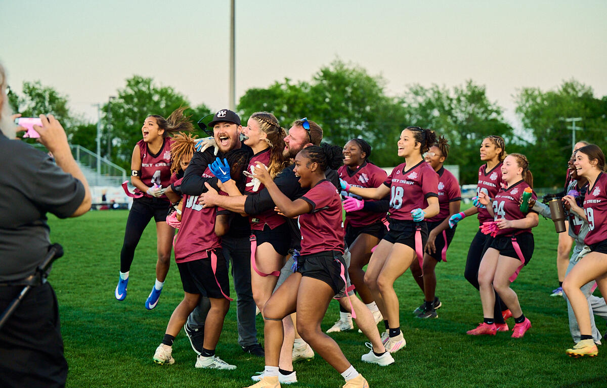 A group of female athletes in maroon uniforms celebrate on a grassy field, hugging and cheering as a person in the foreground records with a pink phone.