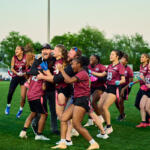 A group of female athletes in maroon uniforms celebrate on a grassy field, hugging and cheering as a person in the foreground records with a pink phone.