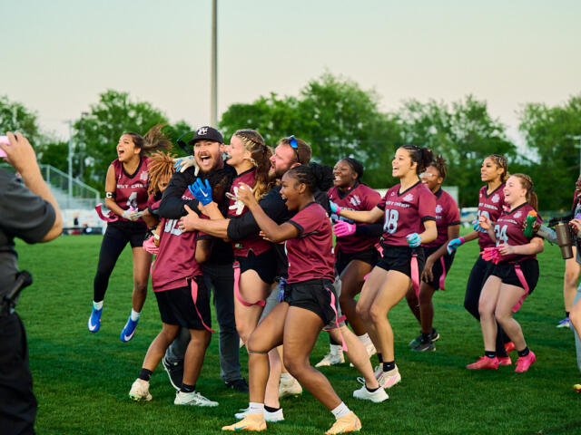 A group of female athletes in maroon uniforms celebrate on a grassy field, hugging and cheering as a person in the foreground records with a pink phone.
