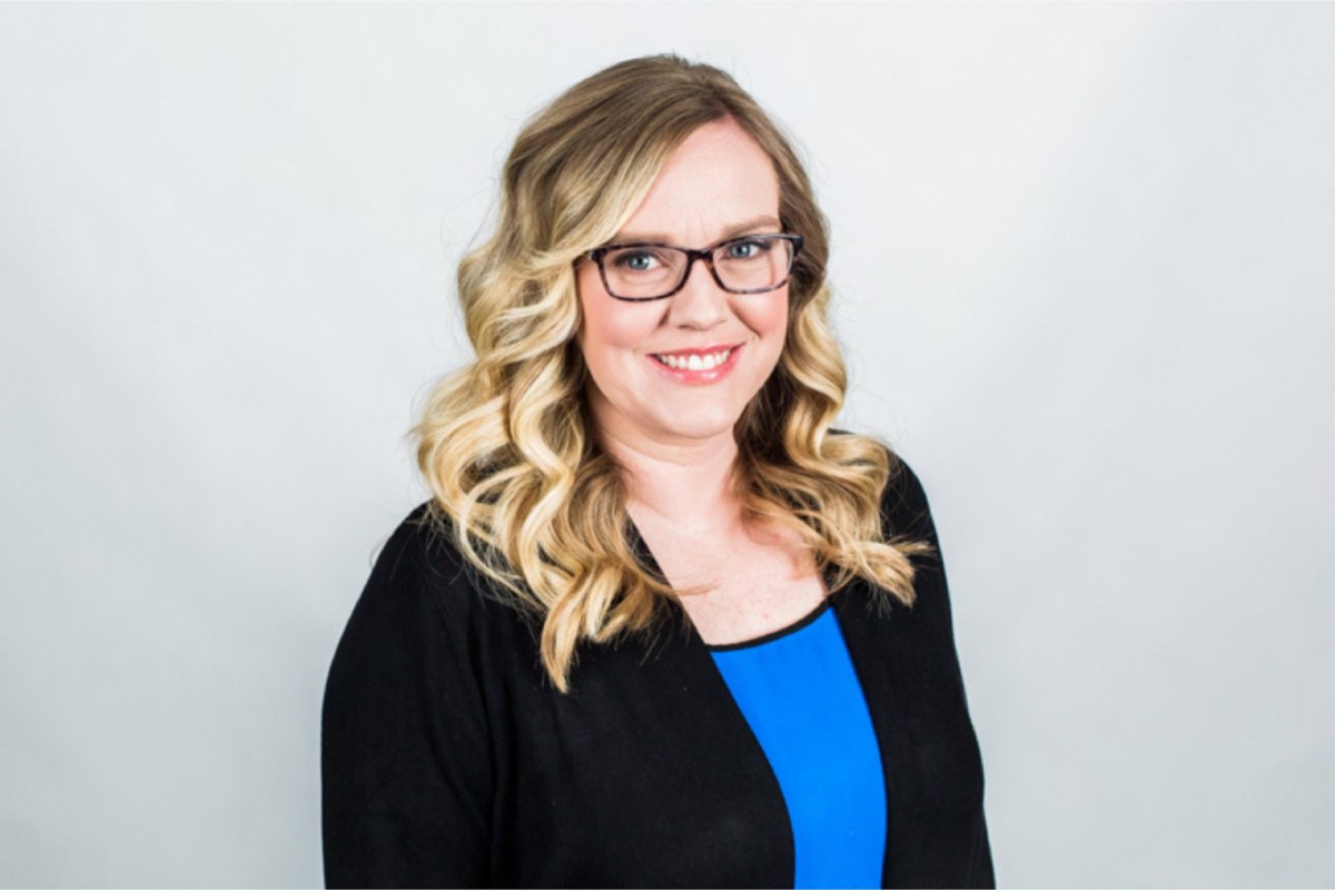 Smiling woman with glasses and blonde curls, wearing a blue top and black cardigan against a light gray background (professional headshot).