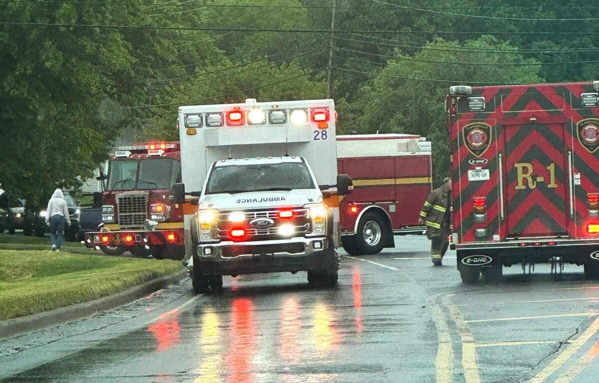 Emergency vehicles including an ambulance and fire trucks line a wet street during rain, with a firefighter approaching the rear of a red truck.