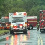 Emergency vehicles including an ambulance and fire trucks line a wet street during rain, with a firefighter approaching the rear of a red truck.