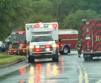 Emergency vehicles including an ambulance and fire trucks line a wet street during rain, with a firefighter approaching the rear of a red truck.