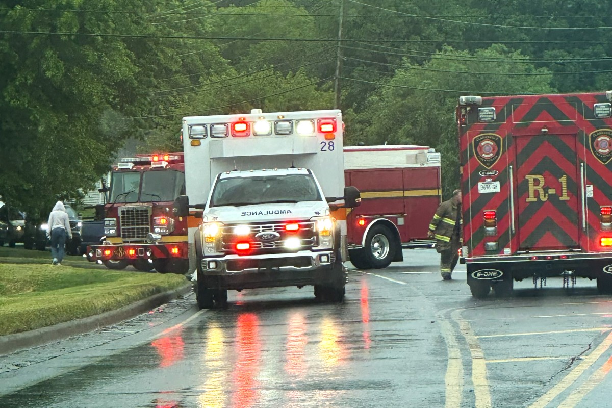 Emergency vehicles including an ambulance and fire trucks line a wet street during rain, with a firefighter approaching the rear of a red truck.