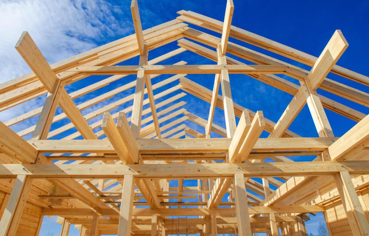Wooden frame of a house under construction against a blue sky with exposed beams and roof trusses, upward perspective