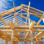 Wooden frame of a house under construction against a blue sky with exposed beams and roof trusses, upward perspective