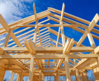 Wooden frame of a house under construction against a blue sky with exposed beams and roof trusses, upward perspective