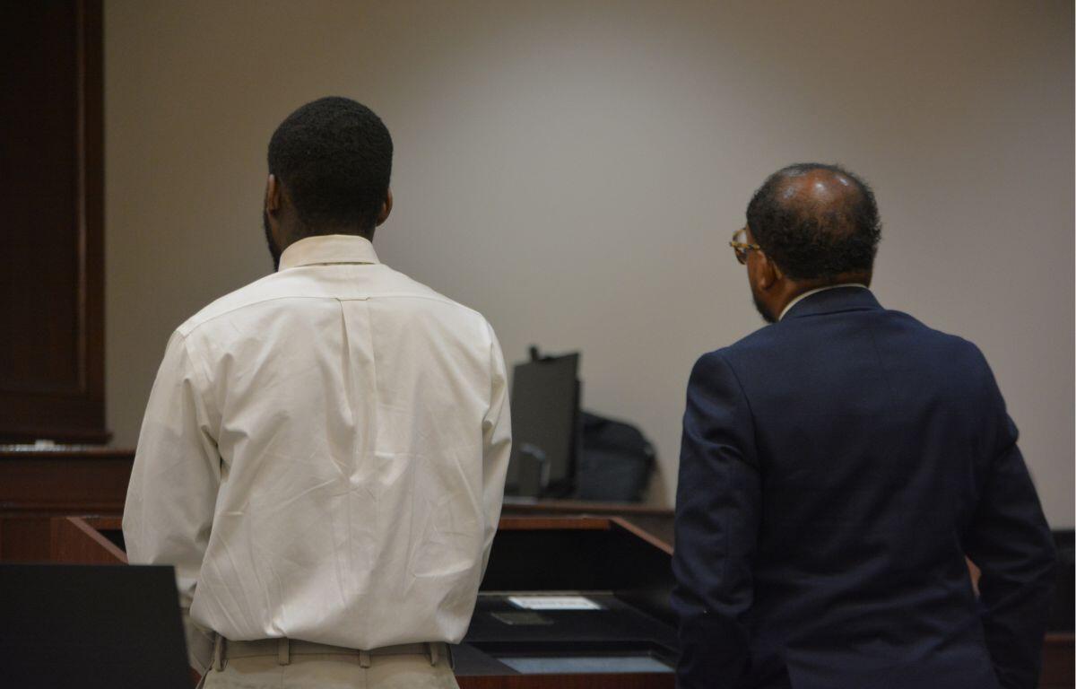 Two men stand with their backs to the camera at a courtroom desk, one in a white shirt and khaki pants and the other in a dark suit.