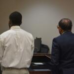 Two men stand with their backs to the camera at a courtroom desk, one in a white shirt and khaki pants and the other in a dark suit.