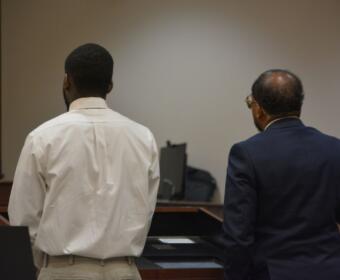Two men stand with their backs to the camera at a courtroom desk, one in a white shirt and khaki pants and the other in a dark suit.