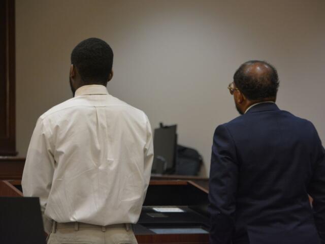 Two men stand with their backs to the camera at a courtroom desk, one in a white shirt and khaki pants and the other in a dark suit.