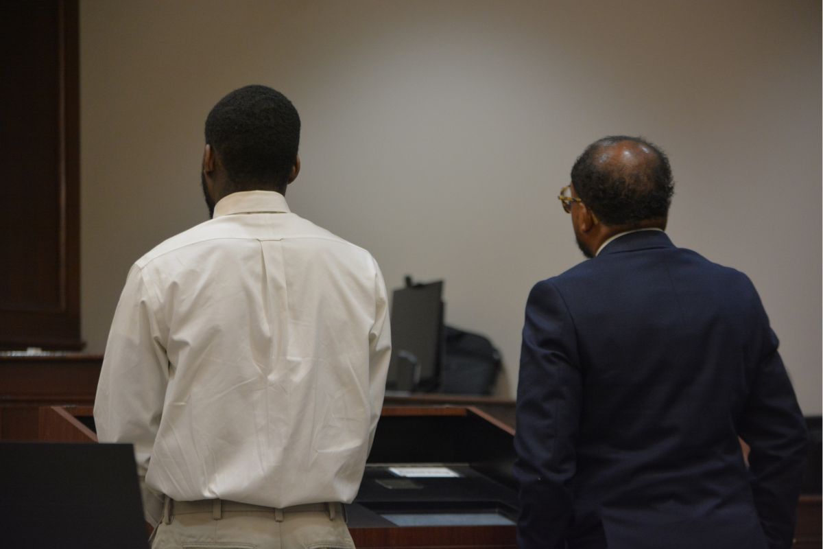 Two men stand with their backs to the camera at a courtroom desk, one in a white shirt and khaki pants and the other in a dark suit.