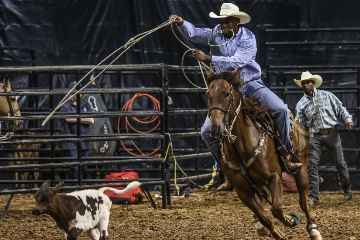 Cowboy on a brown horse swinging a lasso at a calf inside a rodeo arena, with two men watching behind the gate.