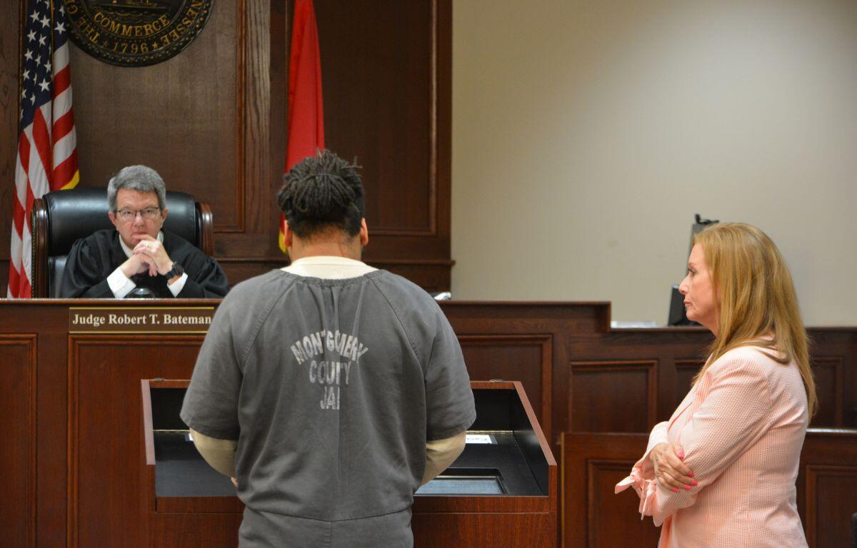 In a courtroom, a judge sits at the bench with hands pressed together, facing a man in a gray inmate jumpsuit who stands with his back to the camera, while a woman in a pink blazer watches from the right.