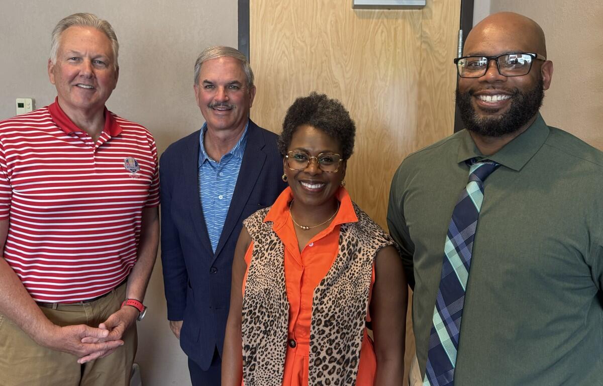 Four adults smiling at the camera posing indoors—a group photo with two men in business-casual attire, a woman in an orange blouse with a leopard-print vest, and a man in a green shirt and tie.