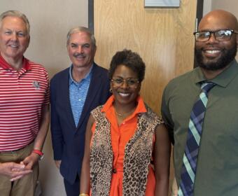 Four adults smiling at the camera posing indoors—a group photo with two men in business-casual attire, a woman in an orange blouse with a leopard-print vest, and a man in a green shirt and tie.