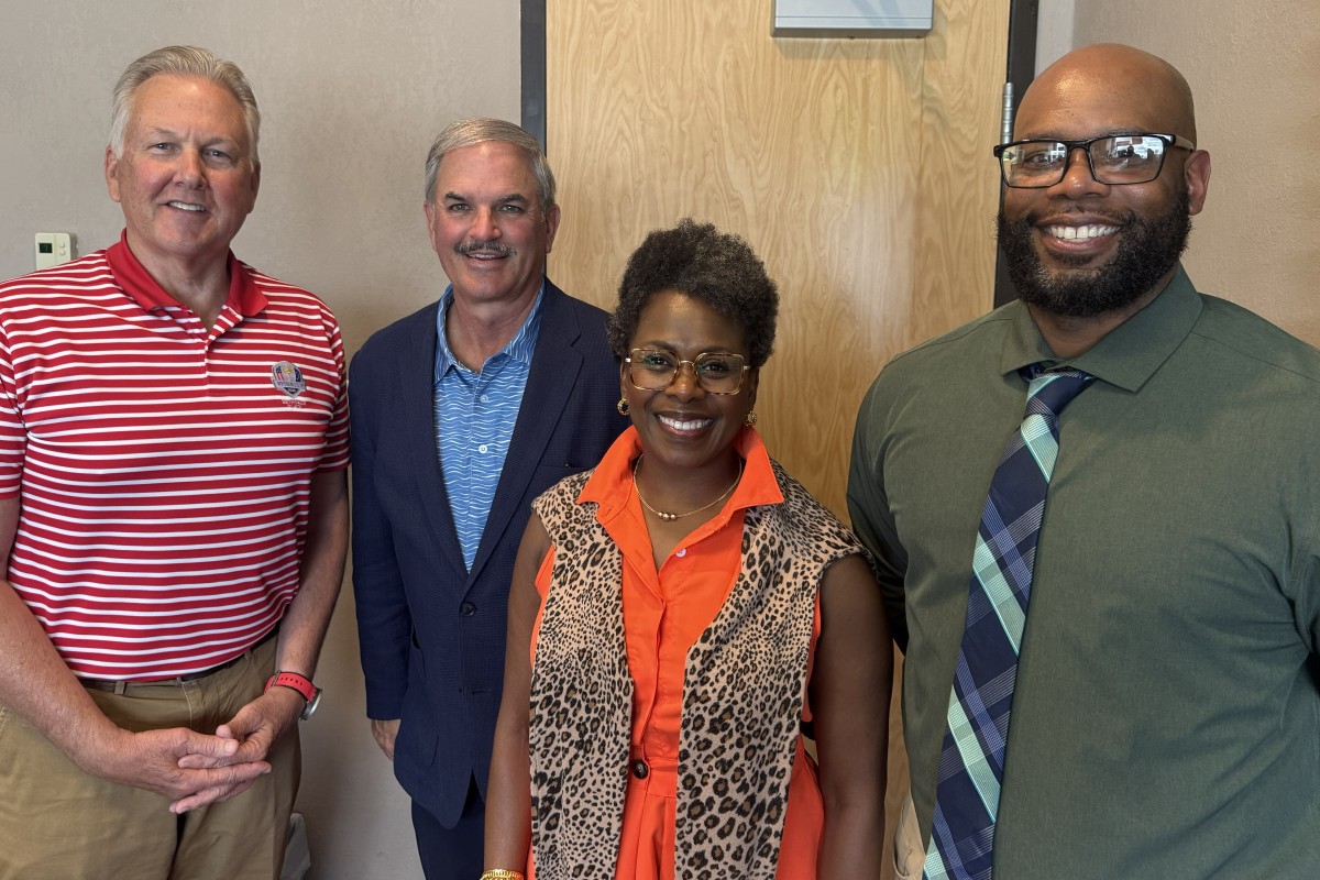 Four adults smiling at the camera posing indoors—a group photo with two men in business-casual attire, a woman in an orange blouse with a leopard-print vest, and a man in a green shirt and tie.