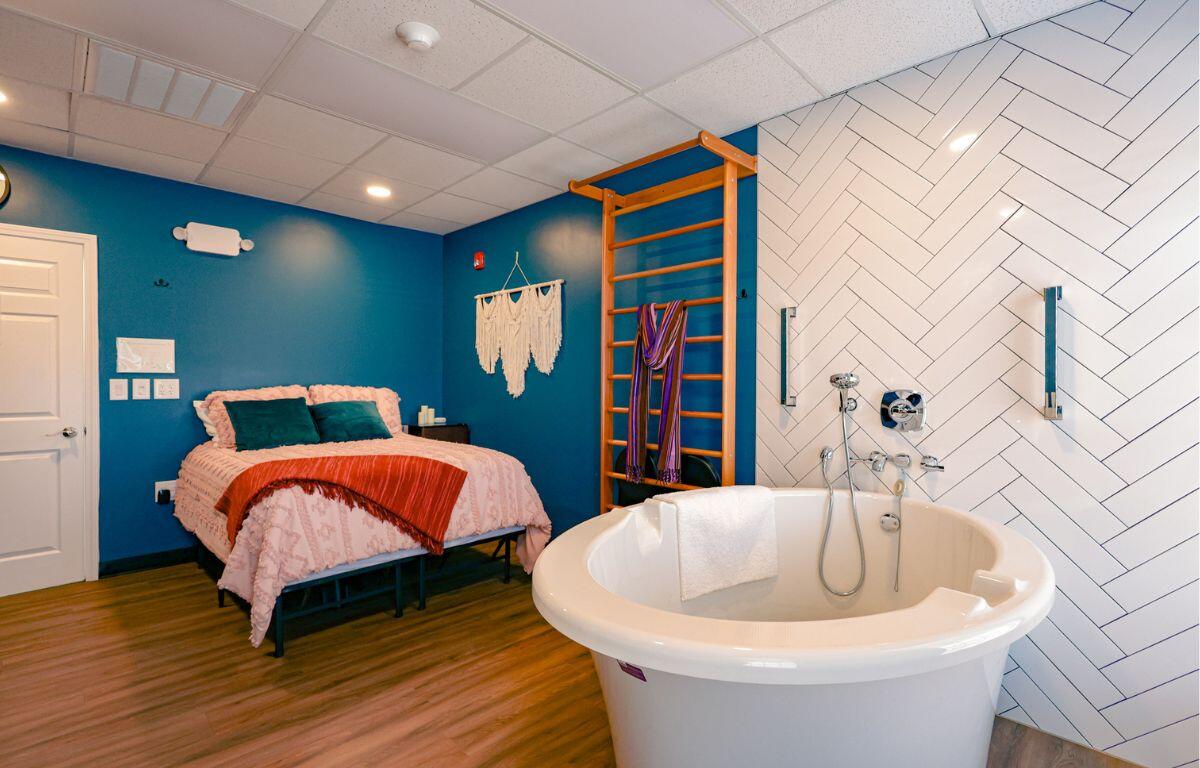 Bedroom-bathroom combo: a freestanding white tub near a blue accent wall, with a pink quilted bed and teal pillows on a wooden floor.