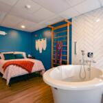 Bedroom-bathroom combo: a freestanding white tub near a blue accent wall, with a pink quilted bed and teal pillows on a wooden floor.