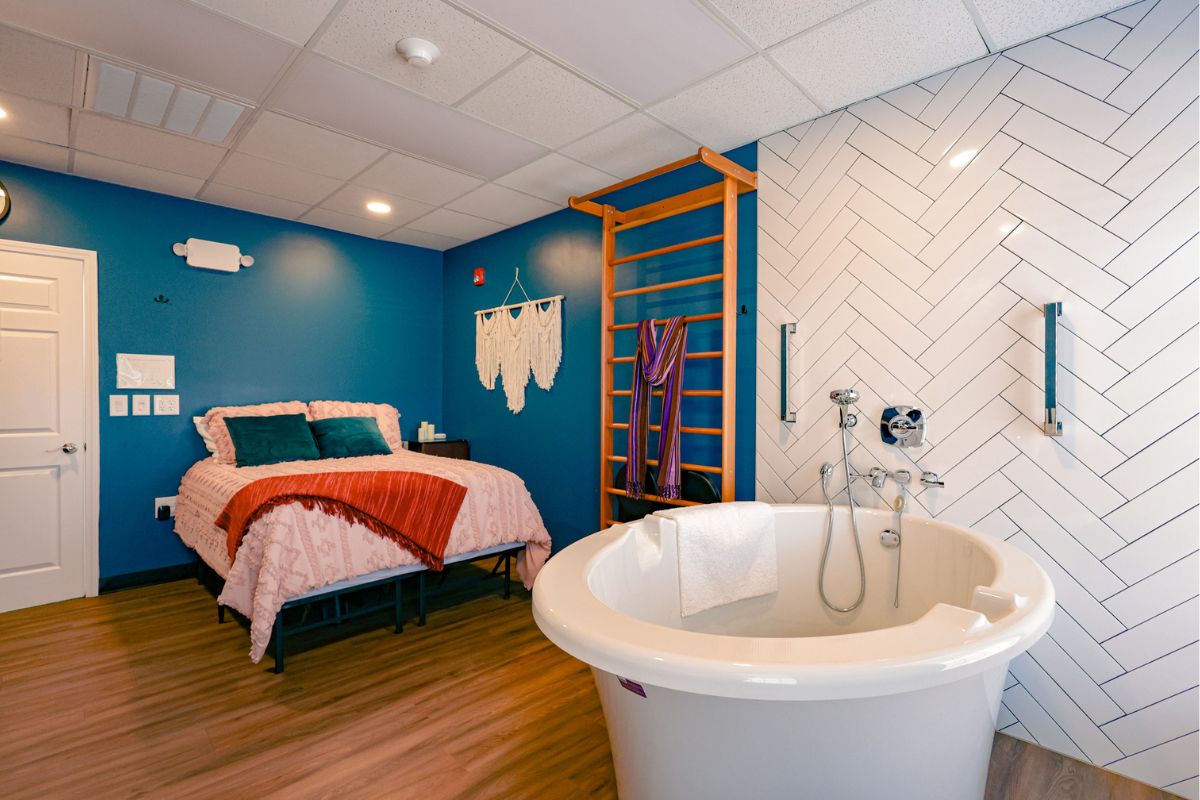 Bedroom-bathroom combo: a freestanding white tub near a blue accent wall, with a pink quilted bed and teal pillows on a wooden floor.