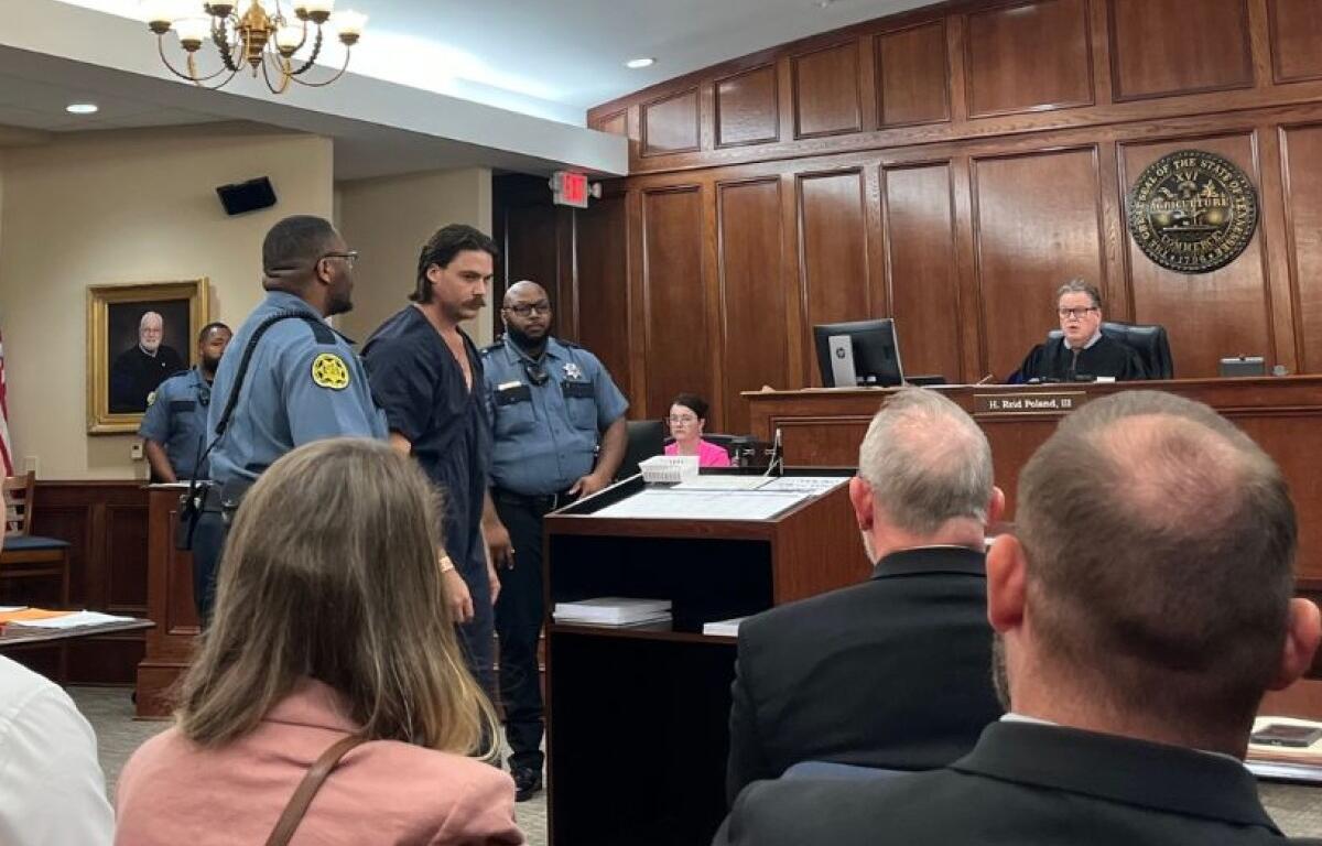 Defendant in a dark jumpsuit escorted by two officers into a courtroom as the judge sits at the bench above them.