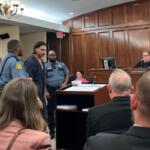 Defendant in a dark jumpsuit escorted by two officers into a courtroom as the judge sits at the bench above them.