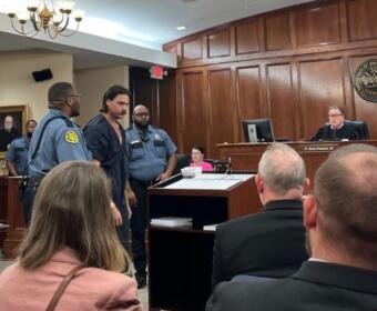 Defendant in a dark jumpsuit escorted by two officers into a courtroom as the judge sits at the bench above them.