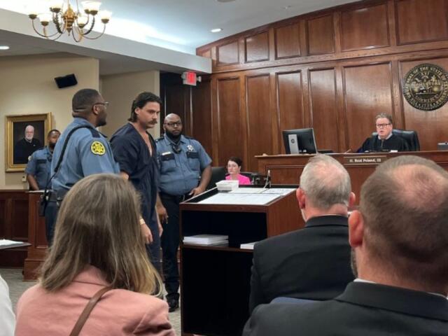 Defendant in a dark jumpsuit escorted by two officers into a courtroom as the judge sits at the bench above them.