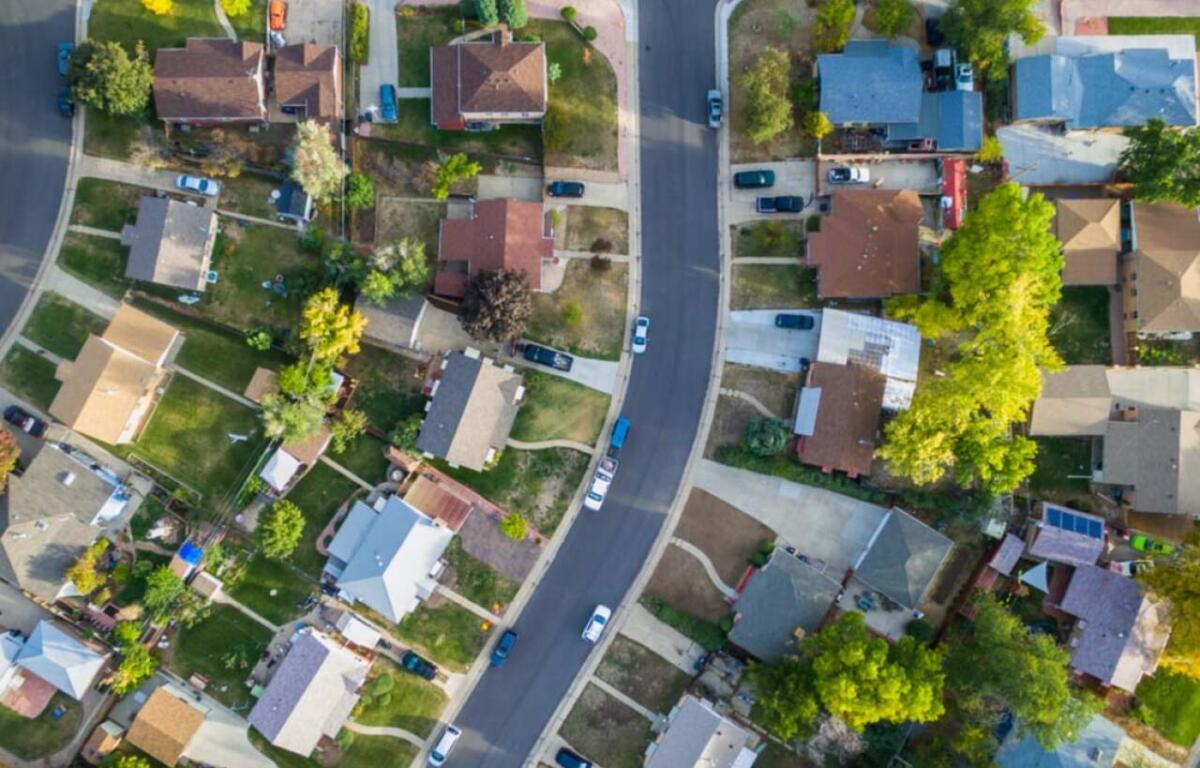 Aerial view of a curved suburban street lined with houses, driveways, cars, lawns, and trees on both sides.