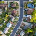 Aerial view of a curved suburban street lined with houses, driveways, cars, lawns, and trees on both sides.
