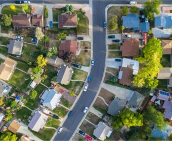 Aerial view of a curved suburban street lined with houses, driveways, cars, lawns, and trees on both sides.