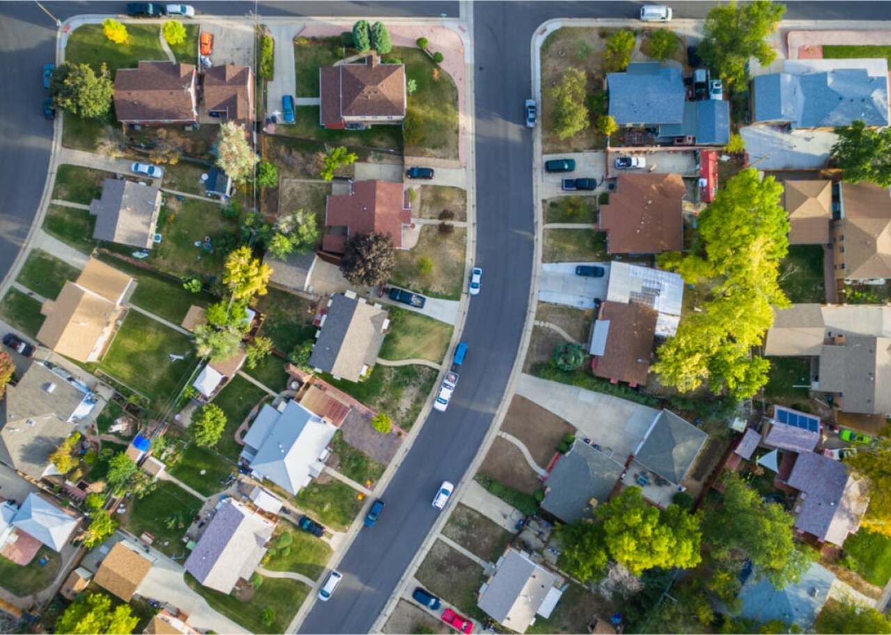 Aerial view of a curved suburban street lined with houses, driveways, cars, lawns, and trees on both sides.