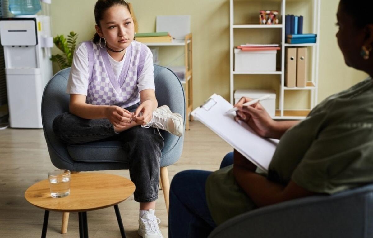 Young woman in a purple argyle vest sits in a chair, talking with a therapist who takes notes on a clipboard in a bright, organized office.