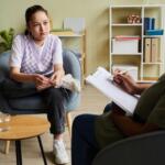 Young woman in a purple argyle vest sits in a chair, talking with a therapist who takes notes on a clipboard in a bright, organized office.