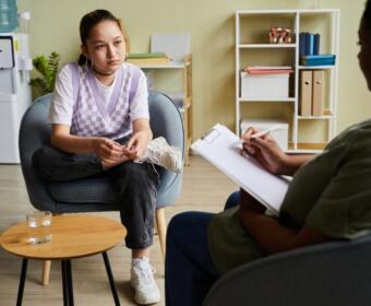 Young woman in a purple argyle vest sits in a chair, talking with a therapist who takes notes on a clipboard in a bright, organized office.