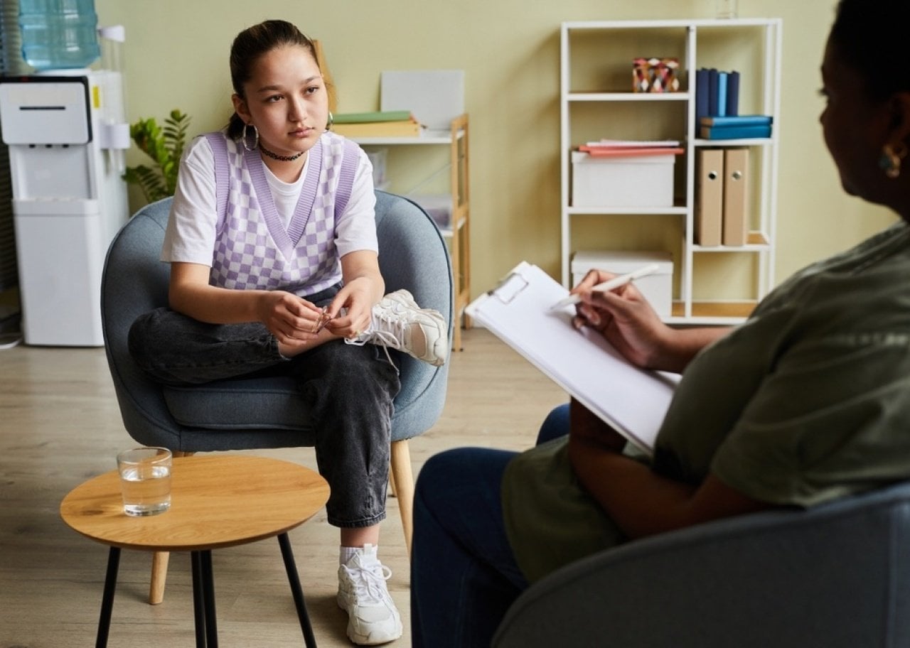 Young woman in a purple argyle vest sits in a chair, talking with a therapist who takes notes on a clipboard in a bright, organized office.