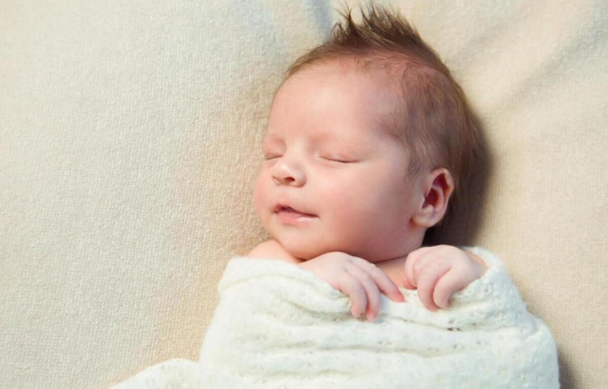 Newborn baby sleeping peacefully, wrapped in a white blanket on a soft beige surface with closed eyes and a relaxed expression.