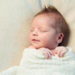 Newborn baby sleeping peacefully, wrapped in a white blanket on a soft beige surface with closed eyes and a relaxed expression.