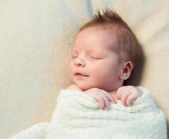 Newborn baby sleeping peacefully, wrapped in a white blanket on a soft beige surface with closed eyes and a relaxed expression.