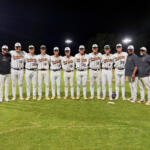A baseball team stands in a line on the field at night for a group photo, wearing white uniforms with maroon writing and caps.